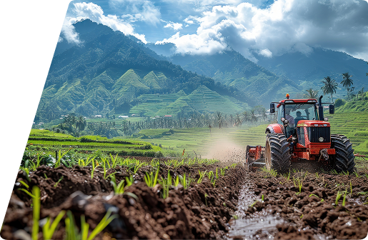A red tractor working in a tilled field with lush green terraced mountains in the background under a cloudy blue sky.