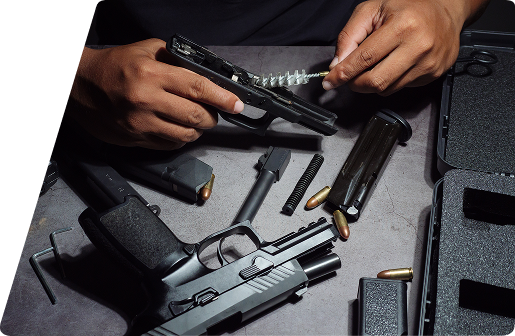 Person cleaning a disassembled handgun with a brush, surrounded by parts, magazines, and ammunition on a work surface.