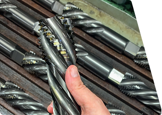 Hand holding a large, metal helical cutting tool or industrial end mill, with other similar tools resting in a rack.